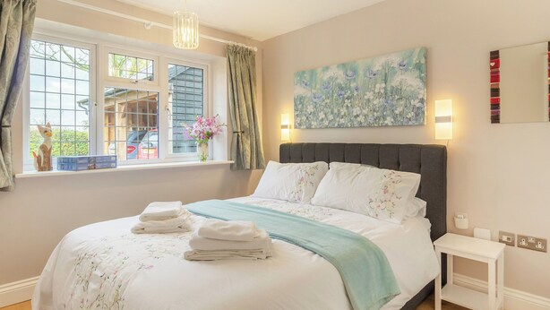 Un dormitorio con cama, ventana con cortinas, un cuadro en la pared y una mesita de noche.
