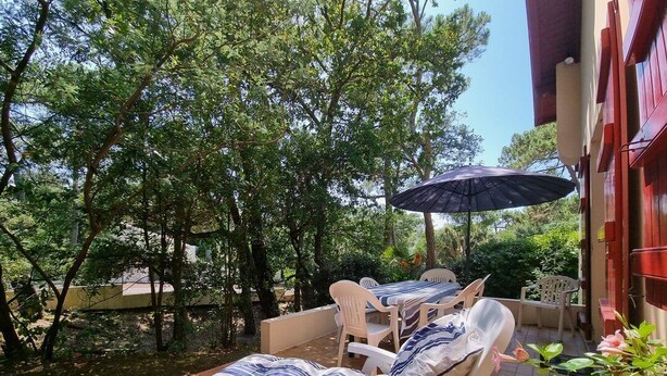 An outdoor seating area with lounge chairs and a table, surrounded by trees and a building with red shutters. - Beach la gravière Villa F4 between Lake and Sea with reversible air conditioning in Hossegor (SOORTS HOSSEGOR)