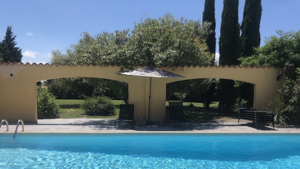 Una piscina cristalina con zona de descanso, que incluye una silla y una sombrilla, rodeada de follaje y árboles altos.