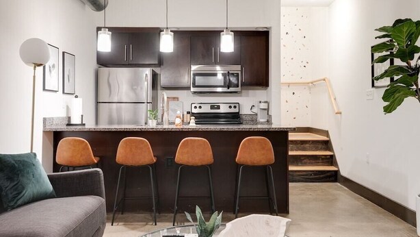 A modern kitchen with dark cabinets, stainless steel appliances, and a bar area with stools. - Atrium 105 - Lovely 1-Bedroom Loft (Richmond)