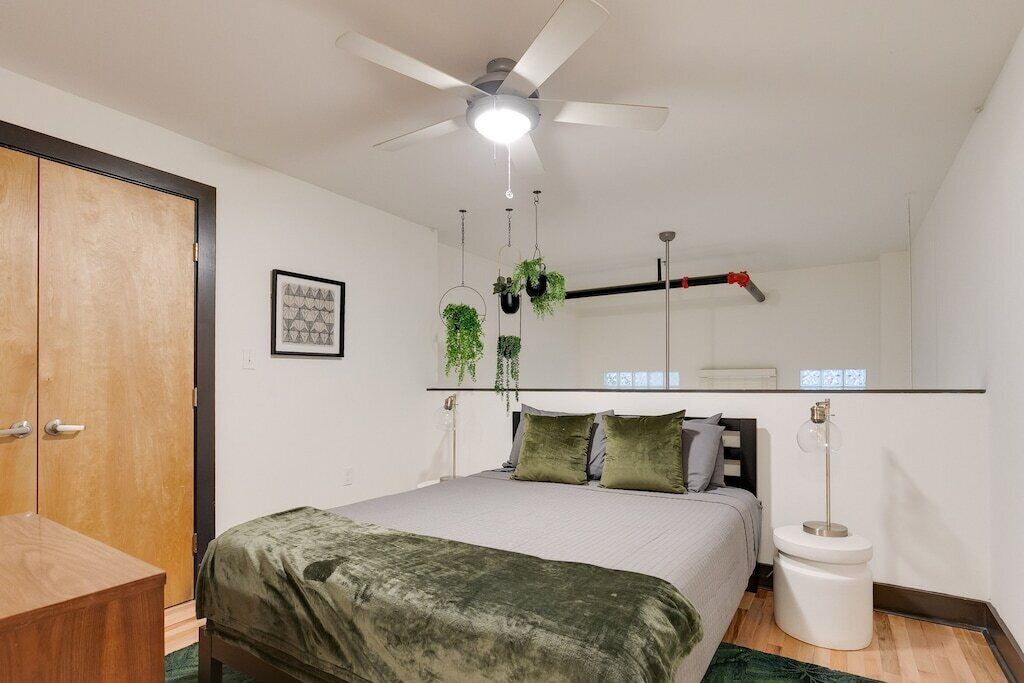A bedroom with a bed, a ceiling fan, a hanging plant, a bedside table, and a toilet.