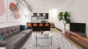 A modern living room with a grey sofa, a coffee table, and a TV. There is a kitchen area with a microwave and a dining set. - Atrium 105 - Lovely 1-Bedroom Loft (Richmond)