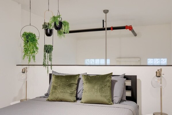 A modern bedroom with a bed, green and grey pillows, hanging plants, and a ceiling-mounted pipe.