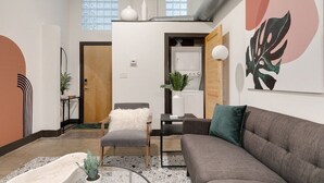 A modern living room with a grey sofa, a white furry ottoman, and a grey armchair. There is a small table with a plant and a white vase. A large abstract painting is on the wall, and a window with blinds is visible. - Atrium 105 - Lovely 1-Bedroom Loft (Richmond)
