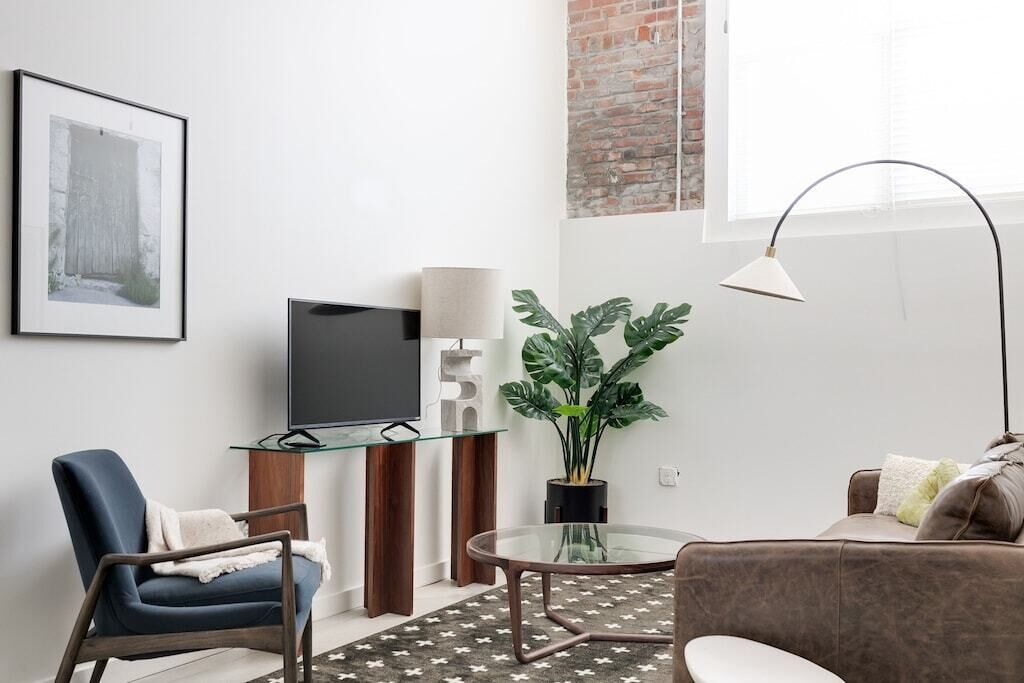 Un salon moderne avec un téléviseur à écran plat, une table basse en verre, un fauteuil bleu, un canapé marron, une grande plante en pot et une lampe sur pied.