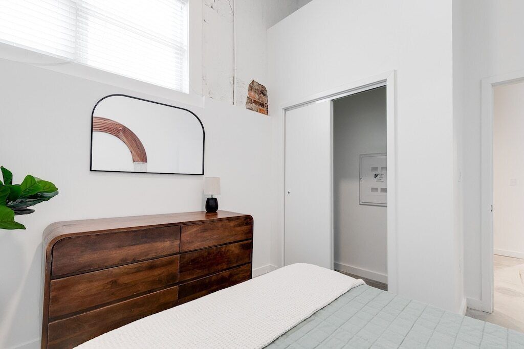 A bedroom with a wooden dresser, a white bed, a mirror, and a door leading to a bathroom.