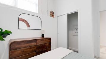 A bedroom with a wooden dresser, a white bed, a mirror, and a door leading to a bathroom.