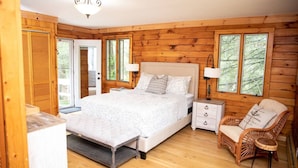 A wooden cabin bedroom with a bed, bedside tables, a chair, and a view of trees through large windows. - Waterfront Home at the  River Haven Sanctuary (Richmond)