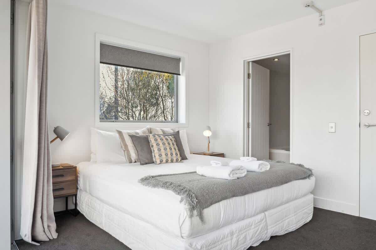 A modern bedroom with a large bed, a window with a view of trees, a bedside table with a lamp, and a door leading to another room.