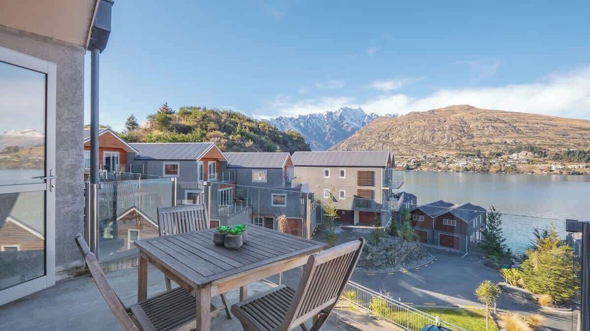 Een balkon met een houten tafel en stoelen, met uitzicht op een meer en bergen.