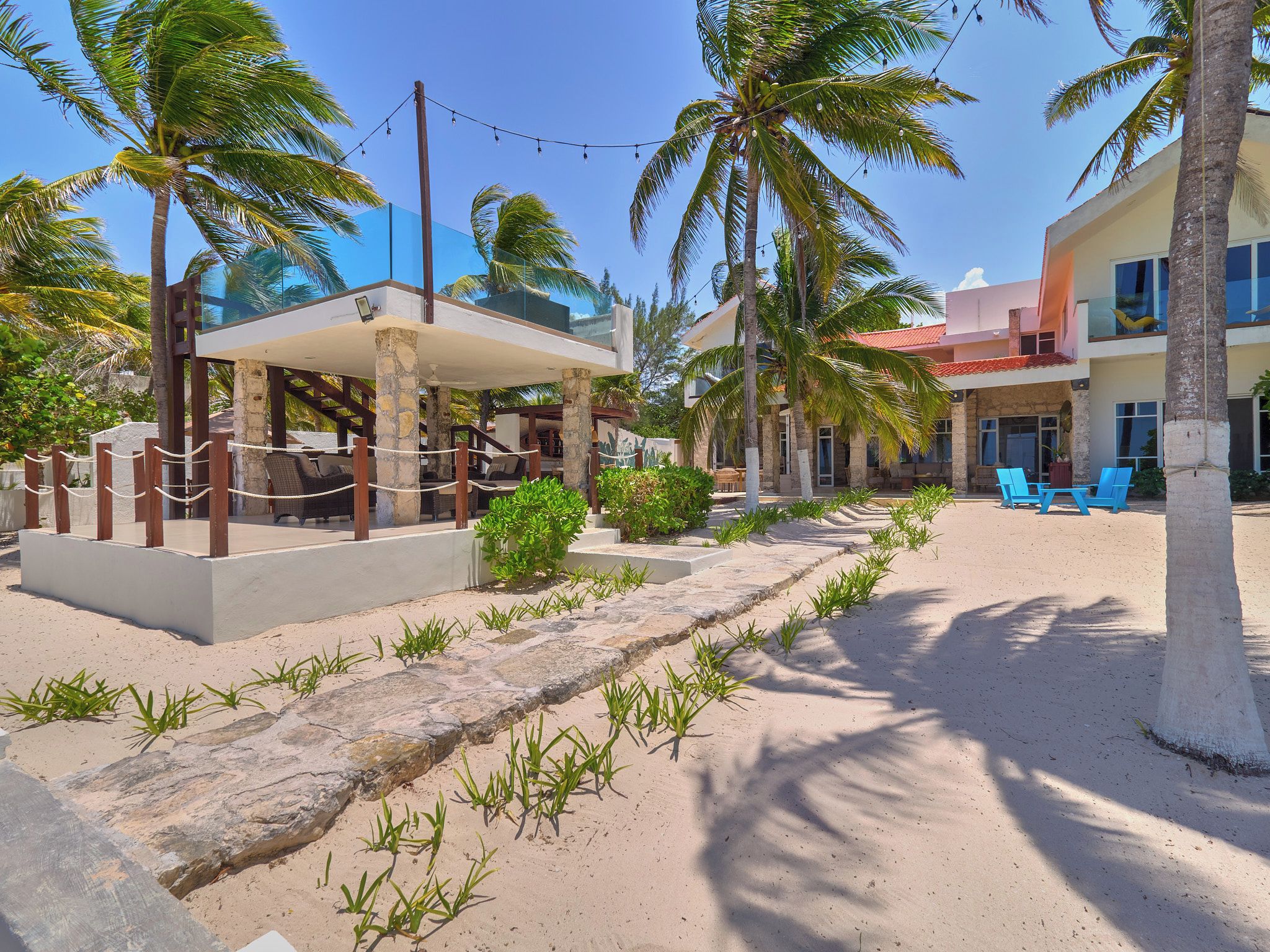 A resort with a covered seating area, palm trees, and a building with blue chairs.