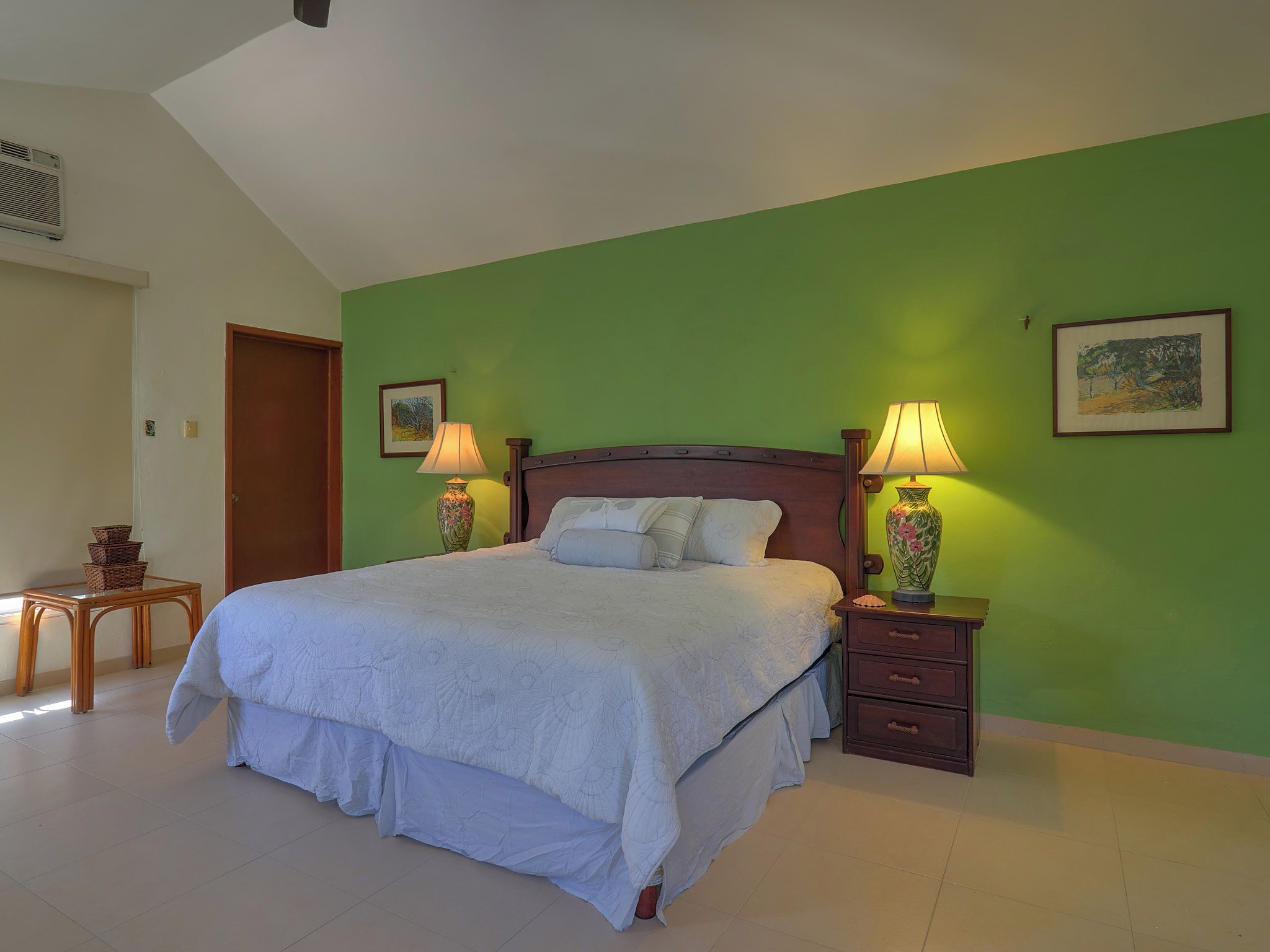 A bedroom with a large bed, two bedside tables with lamps, a small wooden table, and a door.