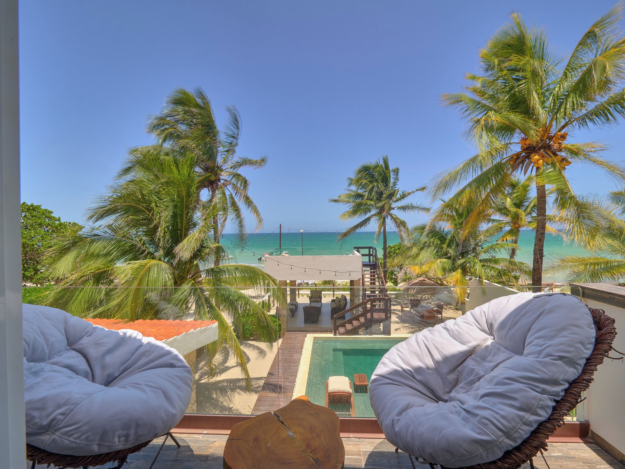 A balcony with two white cushioned chairs, a wooden table, and a view of the ocean and palm trees.