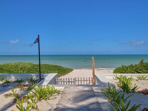 Een strandpad dat naar de oceaan leidt, met een vlag, een houten hek en groene planten.