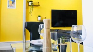Un coin repas avec une table en verre, deux verres à vin, une carafe et un téléviseur en arrière-plan.