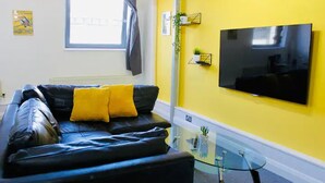 Un salon avec un canapé en cuir noir, deux coussins jaunes, une table basse en verre et un téléviseur à écran plat fixé à un mur jaune.