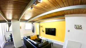 Un salon avec un canapé noir, un téléviseur à écran plat fixé au mur et un mur d’accent jaune.