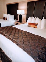 A hotel room with two beds, a patterned bed runner, a nightstand with a lamp, and a headboard with multiple pillows.