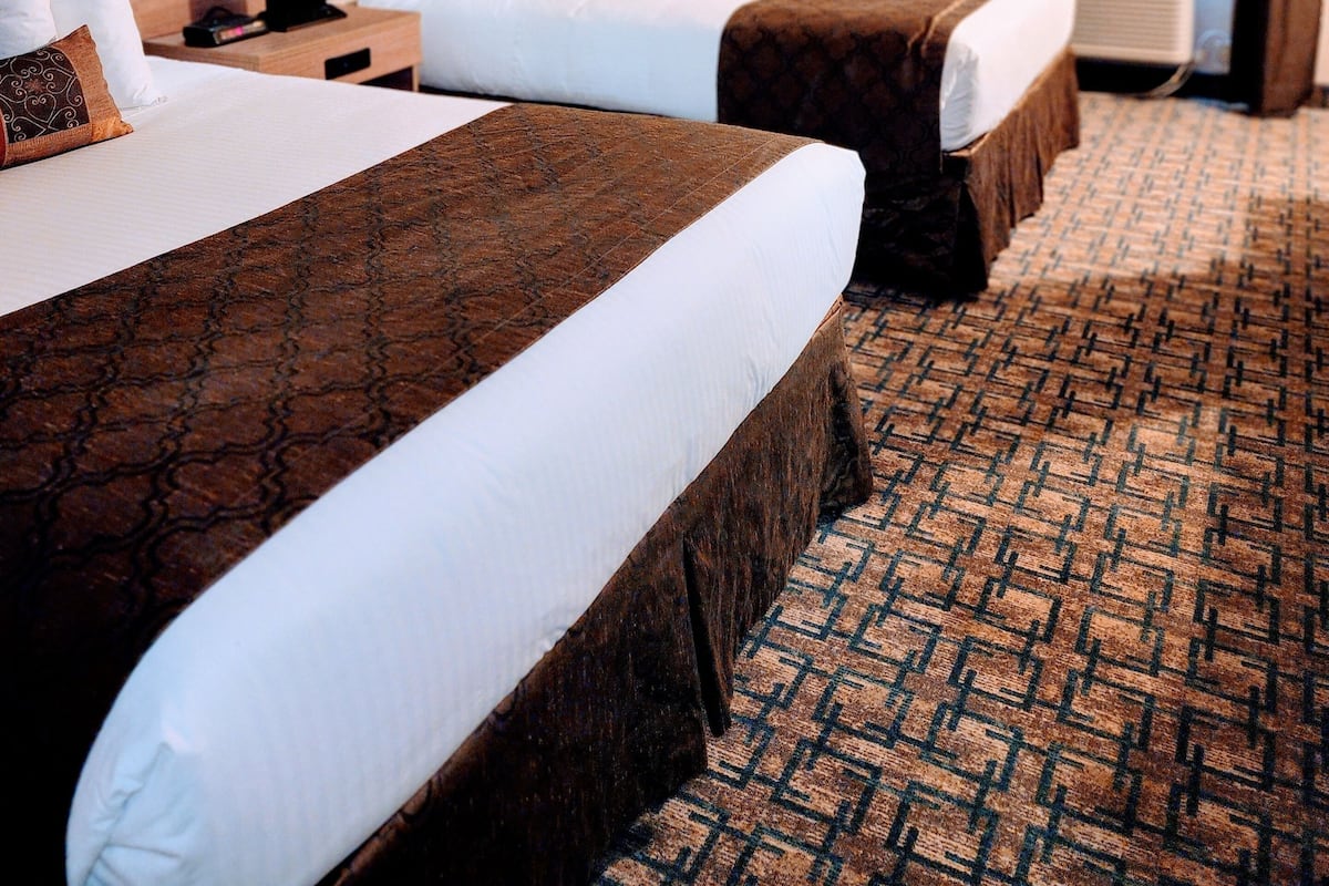 A hotel room with two beds, a nightstand with a lamp, a headboard, a window with curtains, and a patterned carpet.