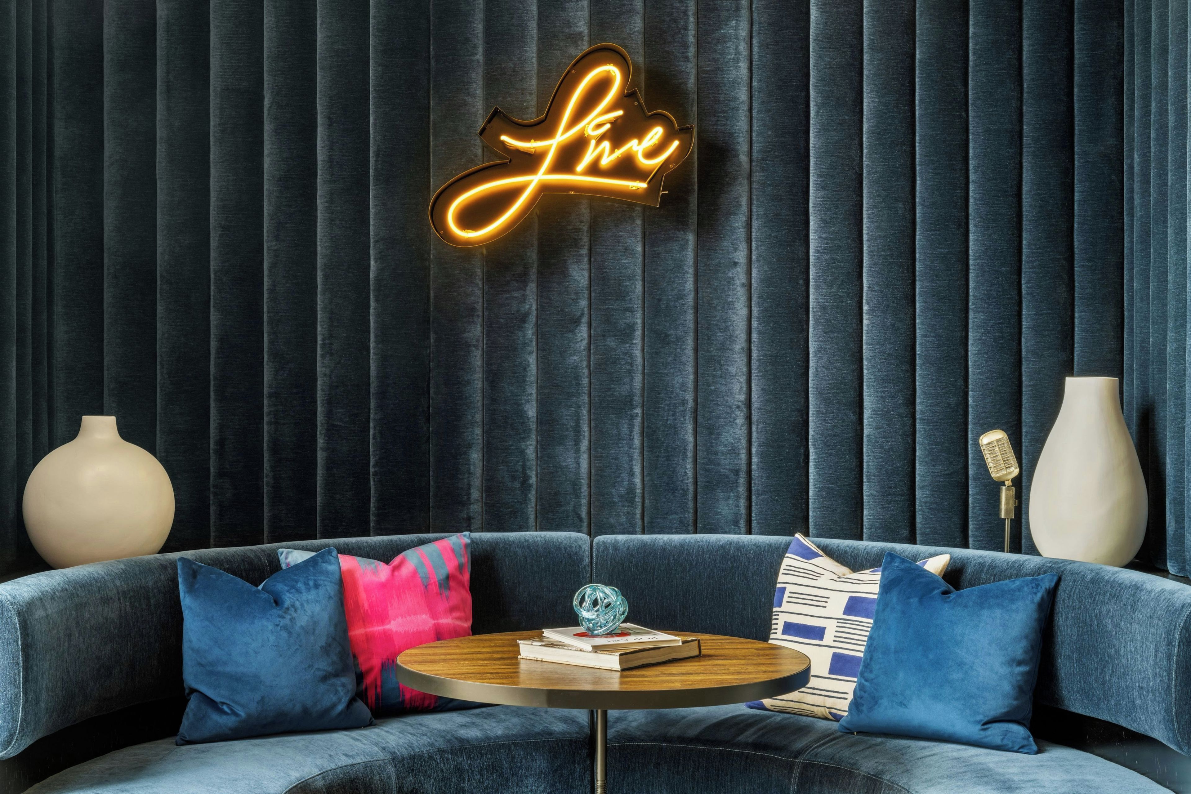 A modern living room with a dark blue velvet draped wall, a neon sign that reads 'love', a round wooden coffee table with books and a glass object, and a blue sofa with patterned cushions.