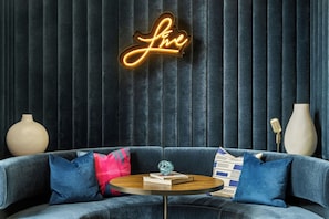 A modern living room with a dark blue velvet draped wall, a neon sign that reads 'love', a round wooden coffee table with books and a glass object, and a blue sofa with patterned cushions. - Aloft by Marriott Chicago Schaumburg (Rolling Meadows)