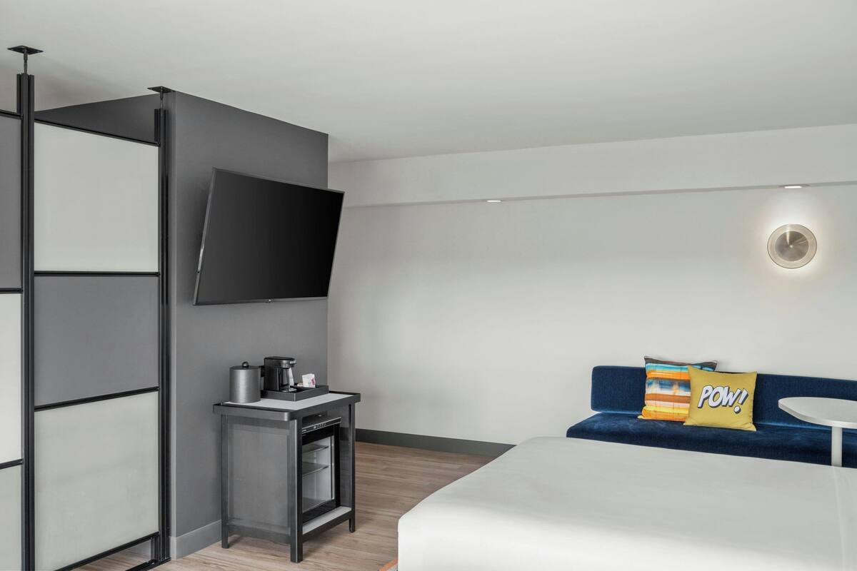 Una habitación de hotel moderna con una cama, un escritorio pequeño con cafetera, un televisor montado en la pared y un sofá azul con dos almohadas.