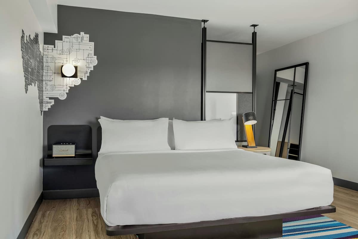 Una habitación de hotel moderna con una cama grande, una mesita de noche con un radio, una lámpara de pared y un espejo.