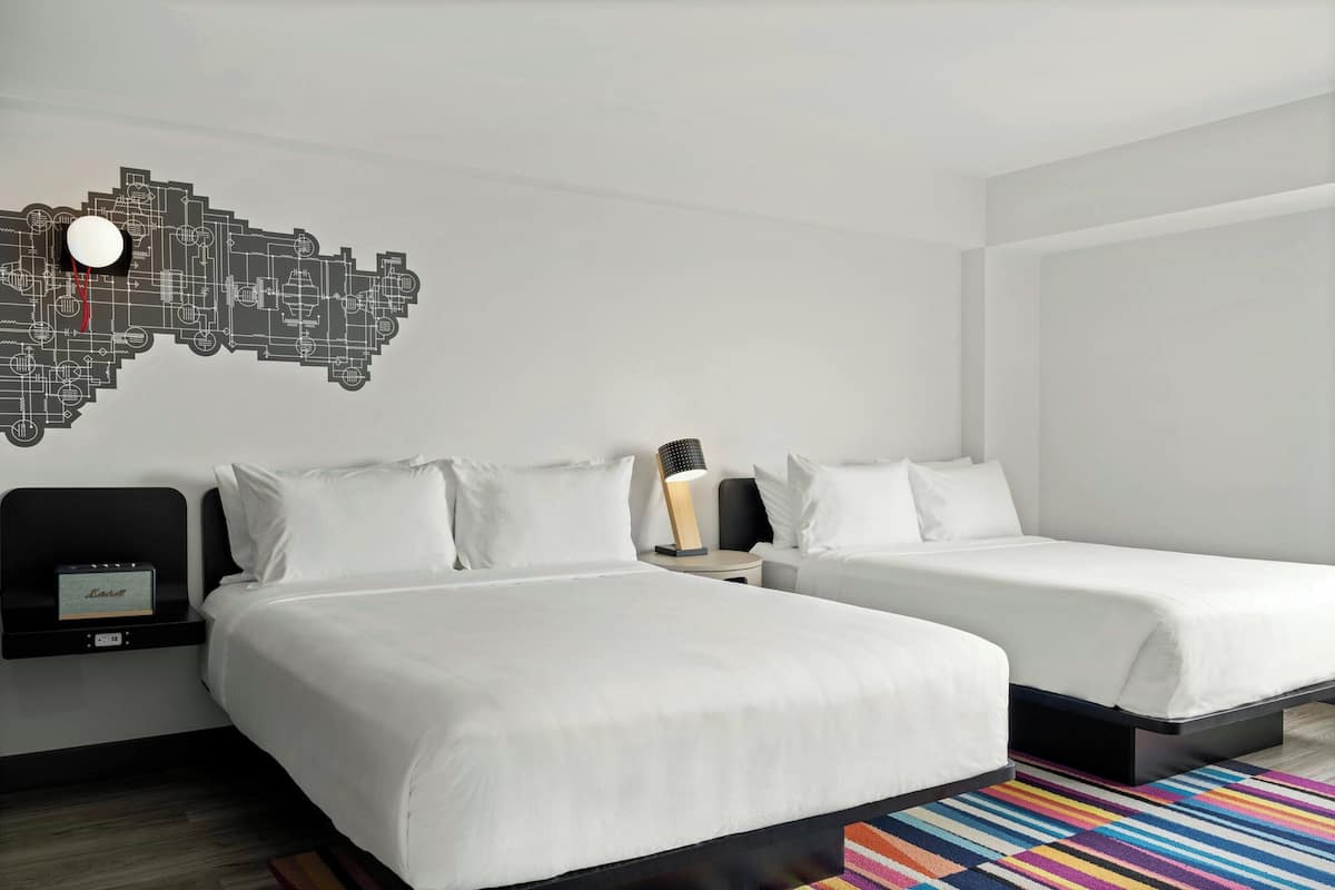 Habitación de hotel con dos camas, una mesita de noche con lámpara, un reloj de pared y una alfombra colorida.