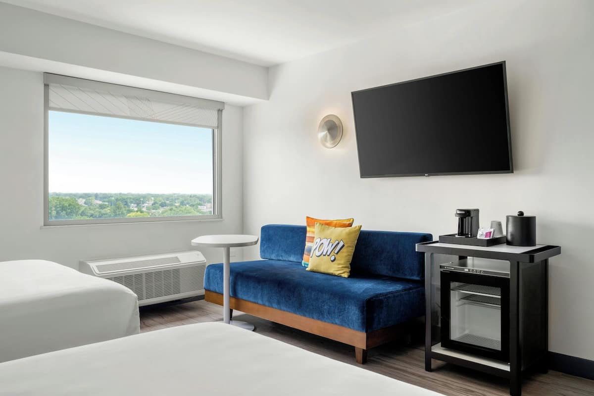 Una habitación de hotel moderna con un sofá azul, un televisor de pantalla plana, una mesita con cafetera y vistas a la ciudad.