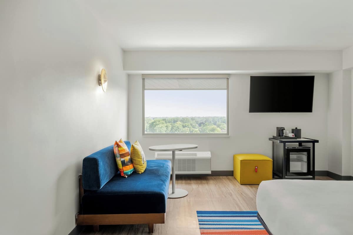 Una habitación de hotel moderna con un sofá azul, una cama, una mesita redonda pequeña, un reposapiés amarillo, un televisor de pantalla plana y un amplio ventanal con vistas a la ciudad.
