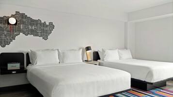 A hotel room with two beds, a wall clock, a side table with a lamp, and a colorful rug.