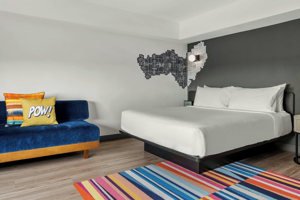 Una habitación de hotel moderna con una cama, un sofá azul, un colorido tapete a rayas y una obra de arte en la pared.