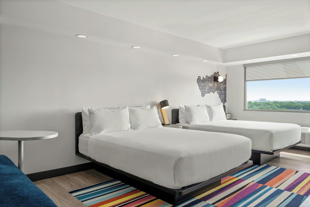 Una habitación de hotel moderna con dos camas, una mesita redonda pequeña, un tapete a rayas de colores y un ventanal con vistas a la ciudad.