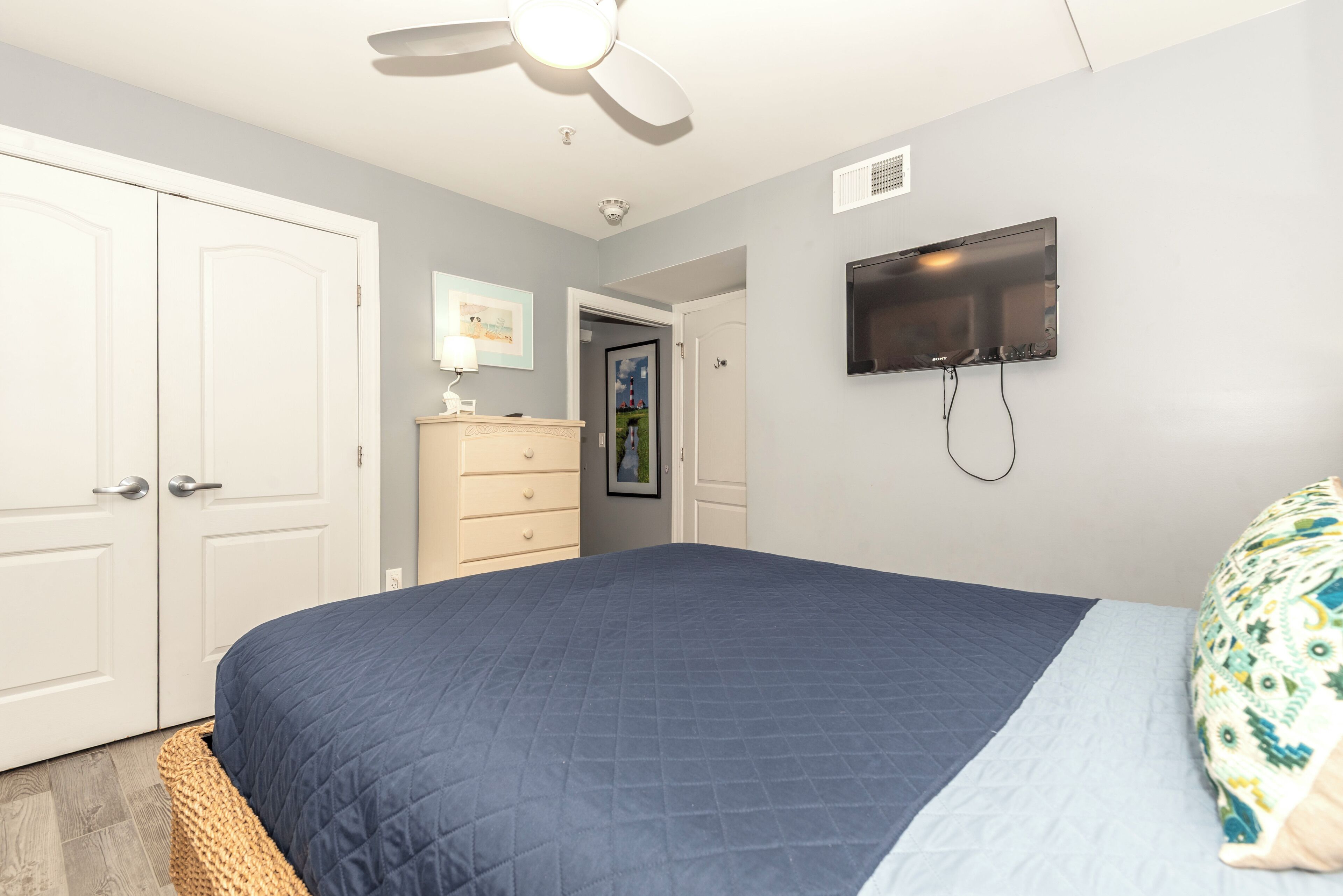 A bedroom with a bed, a ceiling fan, a TV, a dresser, and a door.