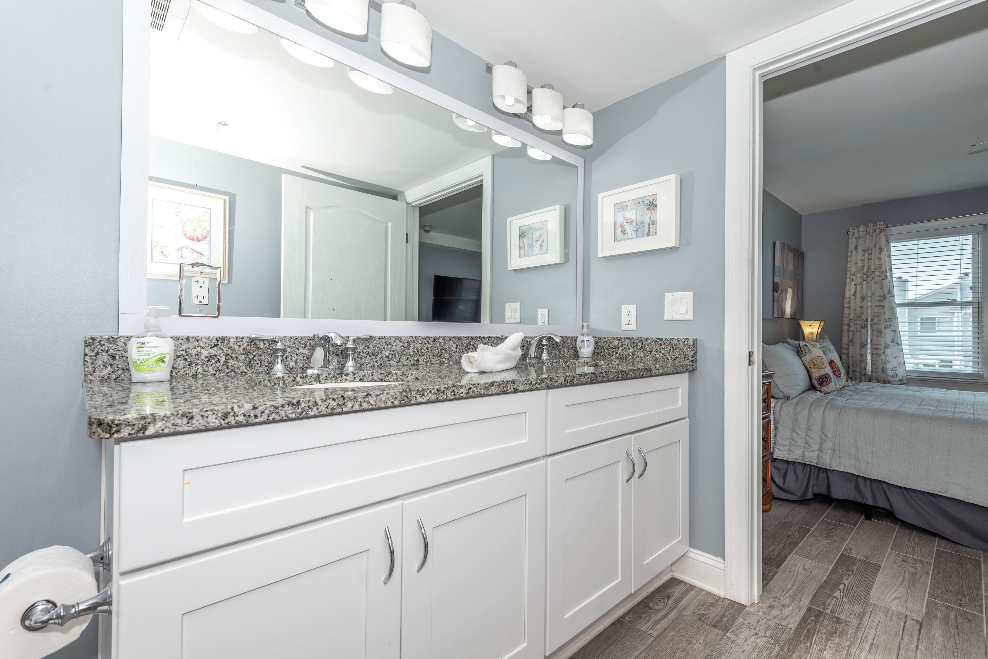 A bathroom with a granite countertop, white cabinets, a large mirror, and a door leading to a bedroom.