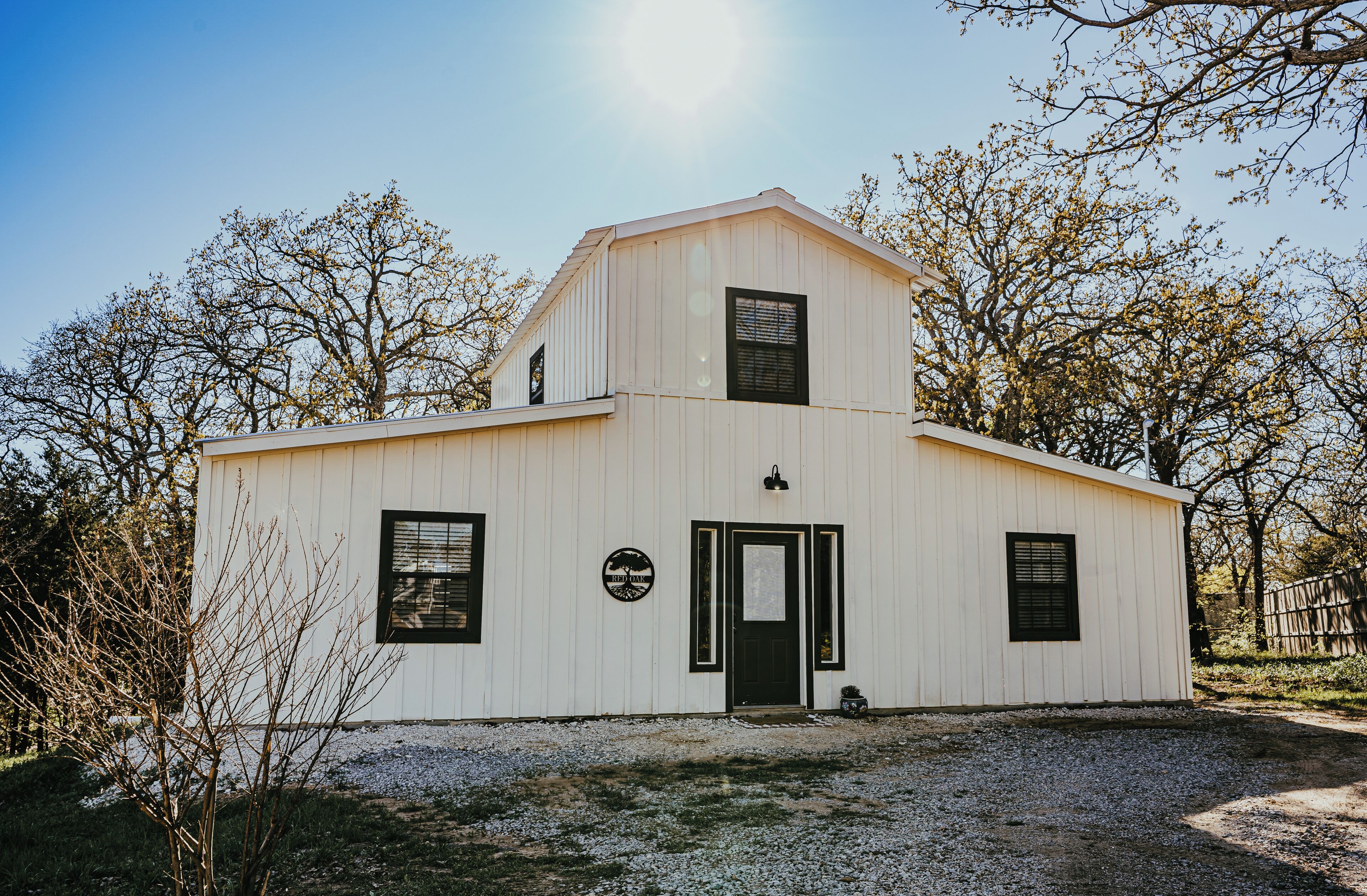 A white building with a black door and windows, surrounded by trees and gravel.