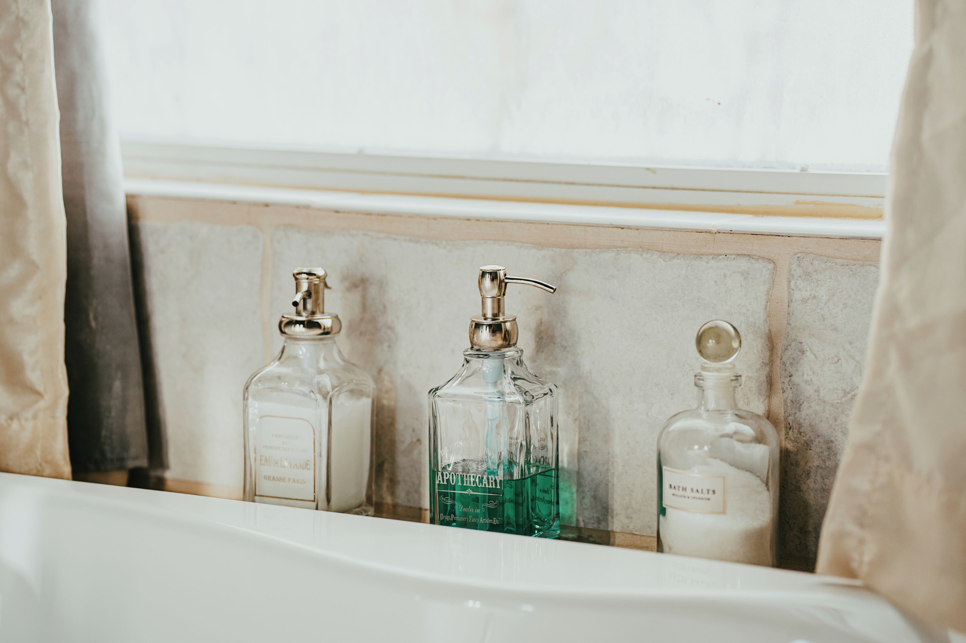 Ett badrum med ett vitt handfat, tre glasflaskor med hygienprodukter och ett fönster med gardiner.