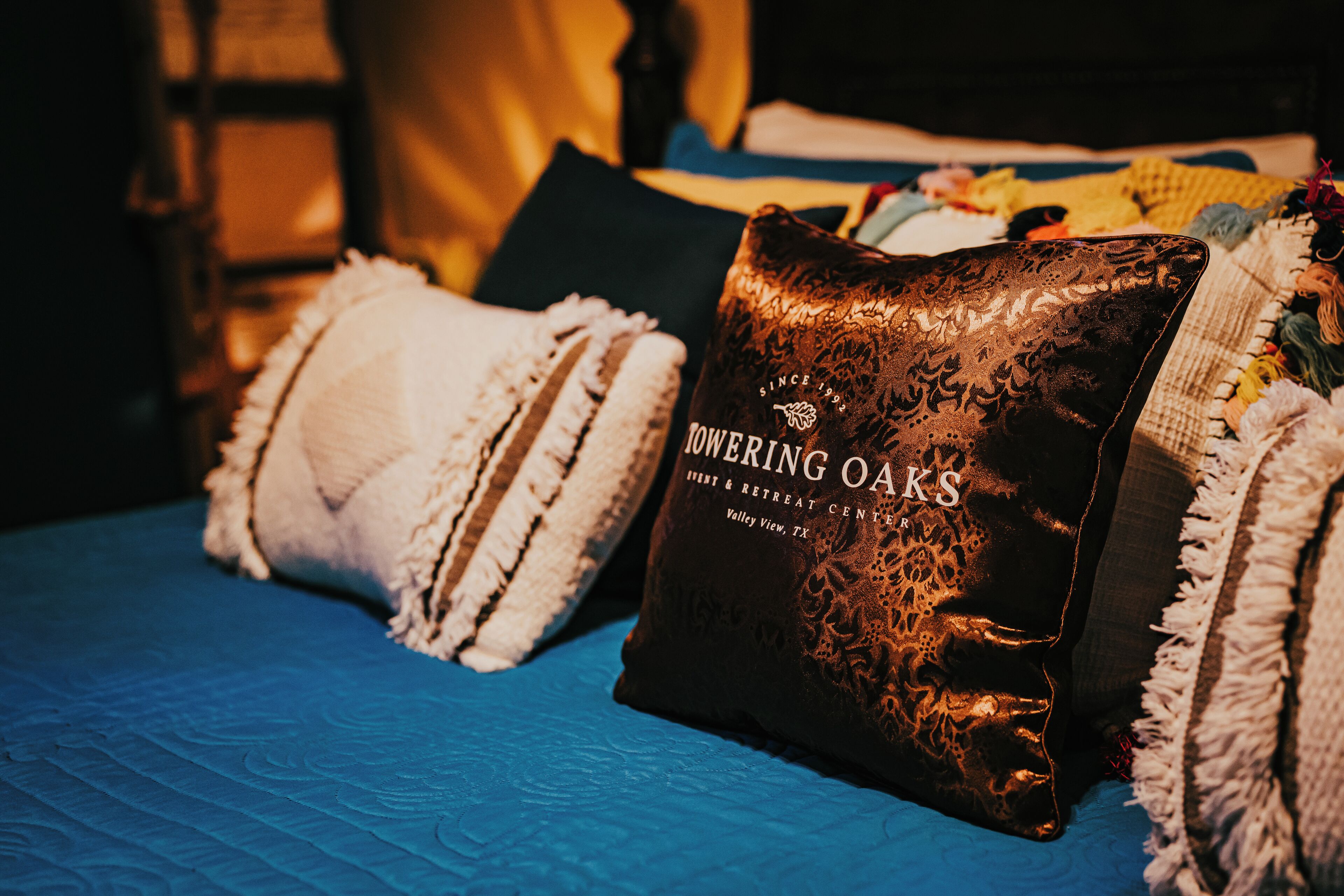 Una cama con colcha azul, dos almohadas, una con estampado floral y la otra con diseño texturizado, y una almohada marrón oscuro con texto en relieve.