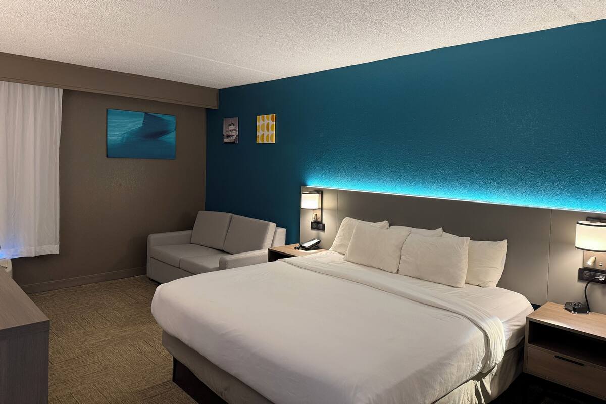 Quarto de hotel com uma cama grande, uma escrivaninha, uma cadeira e uma mesinha de cabeceira.