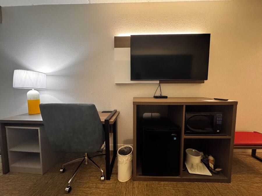 A hotel room with a desk, chair, TV, microwave, and a small trash bin.