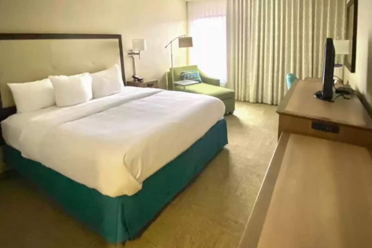 Quarto de hotel com uma cama grande, uma escrivaninha com computador, uma cadeira e uma janela com cortinas.