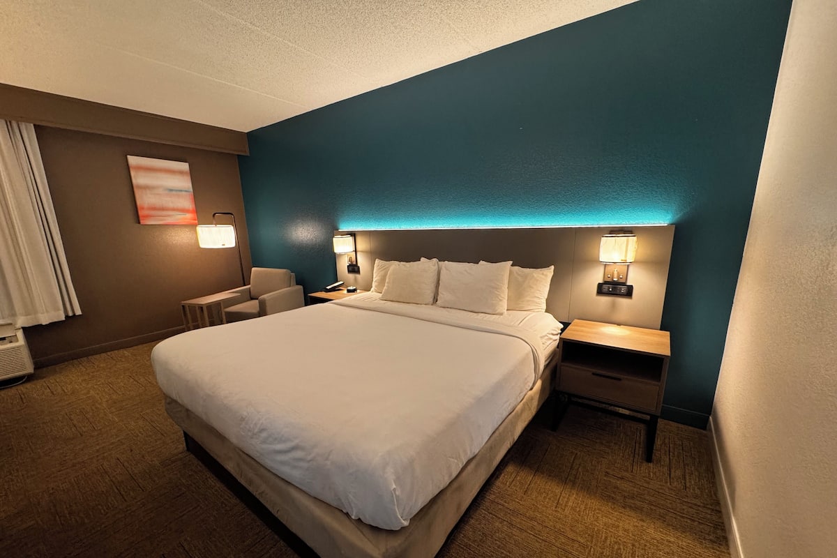 Quarto de hotel com uma cama grande, duas mesas de cabeceira com luminárias, uma cadeira e uma obra de arte na parede.