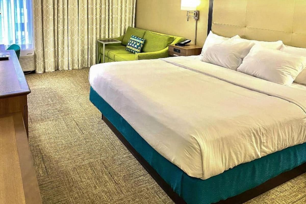 Quarto de hotel com uma cama grande, uma poltrona verde, uma escrivaninha e uma luminária.