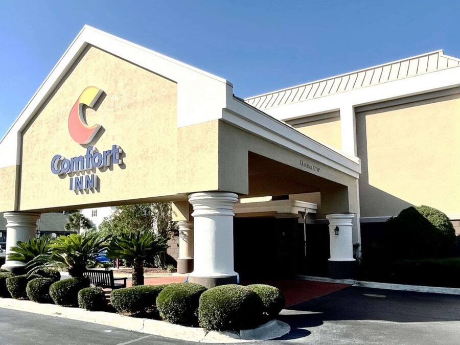 Un hotel Comfort Inn con un logo 'C' destacado, un acceso cubierto y un estacionamiento con arbustos bien cuidados.