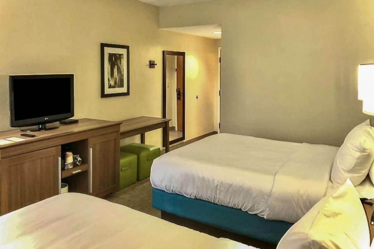 Quarto de hotel com duas camas, uma escrivaninha com televisão, um espelho e uma porta que dá para outro cômodo.