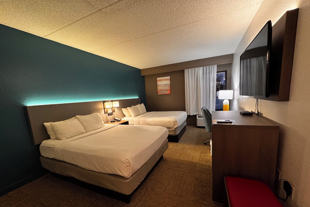 Quarto de hotel com duas camas, uma escrivaninha, uma cadeira, televisão e janela com cortinas.