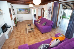 A living room with a purple sofa, a coffee table, and a TV on a stand. There is a painting on the wall and a window with curtains.