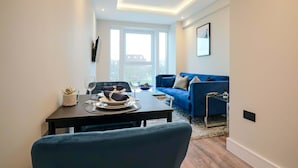 Ein modernes Wohnzimmer mit einem blauen Sofa, einem Esstisch mit Tellern und Gläsern sowie einem großen Fenster, das natürliches Licht hereinlässt.