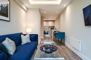 A modern living room with a blue sofa, a glass coffee table with decorative items, and a view into the kitchen area.