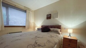 A bedroom with a bed, a nightstand, a lamp, a window with blinds, and a wall-mounted artwork.